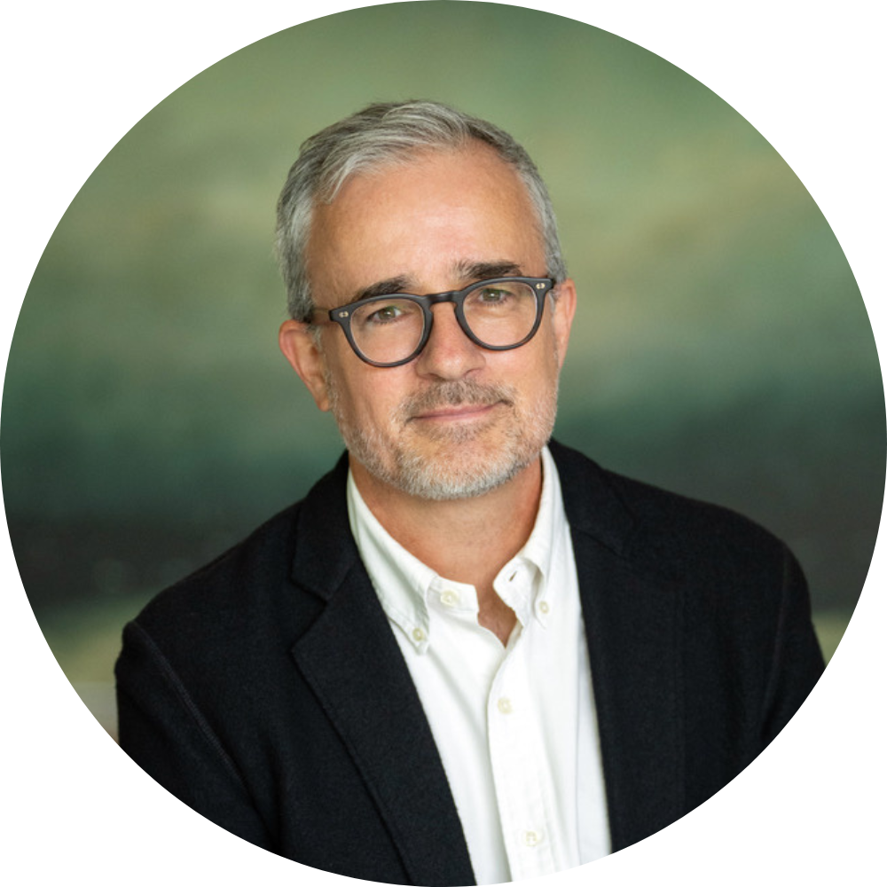 White male with short grey hair and glasses wearing a black blazer and white button-down shirt, seated in front of a green backdrop.