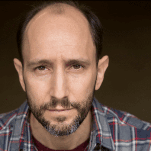 A middle-aged man with a receding hairline and short beard wearing a plaid shirt, looking directly at the camera with a neutral expression.