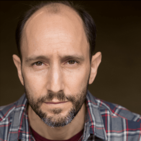 A middle-aged man with a receding hairline and short beard wearing a plaid shirt, looking directly at the camera with a neutral expression.