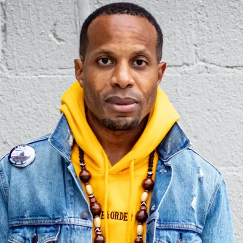 A man wearing a yellow hoodie, denim jacket, and beaded necklace stands against a textured light gray wall.