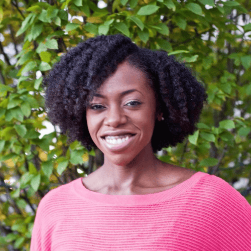 Woman with natural curly hair wearing a pink sweater, smiling in front of green leafy bushes outdoors.