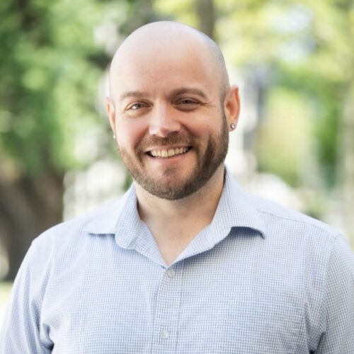 A smiling man with a shaved head and a beard, wearing a light blue button-down shirt.