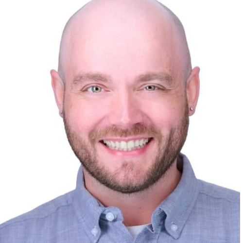 A bald man with a trimmed beard smiles at the camera, wearing a light blue button-up shirt against a white background.