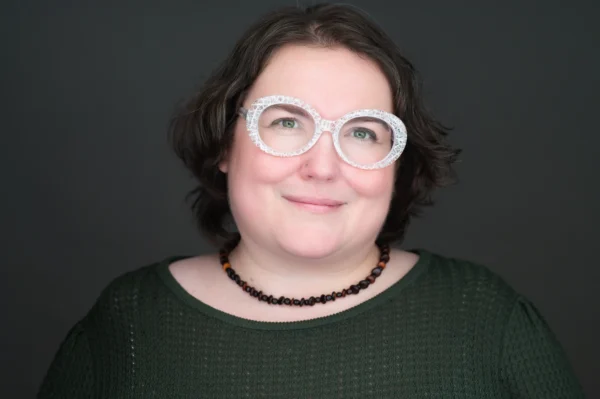 A person with short brown hair wears large round glasses, a dark green textured top, and a beaded necklace, smiling softly against a dark background.