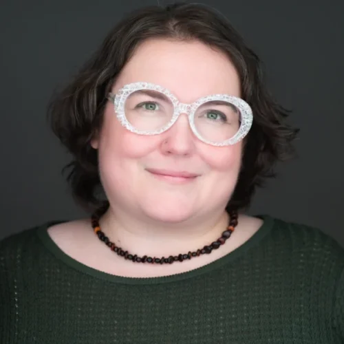A person with short brown hair wears large round glasses, a dark green textured top, and a beaded necklace, smiling softly against a dark background.