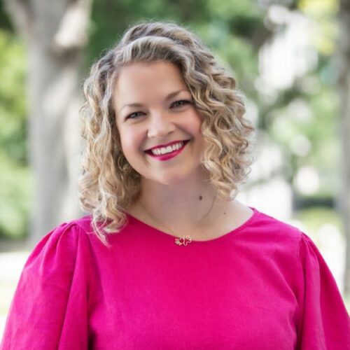 A smiling woman with shoulder-length curly hair wearing a bright pink top and a gold pendant necklace.