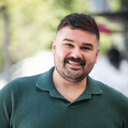 A man with short dark hair and a mustache, wearing a green polo shirt, smiles outdoors with a blurred background of greenery and a building.