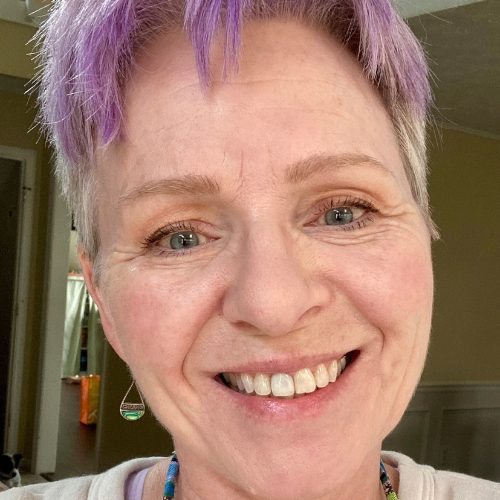 A person with short, light purple hair smiles at the camera indoors, wearing a beaded necklace and a dangling earring.