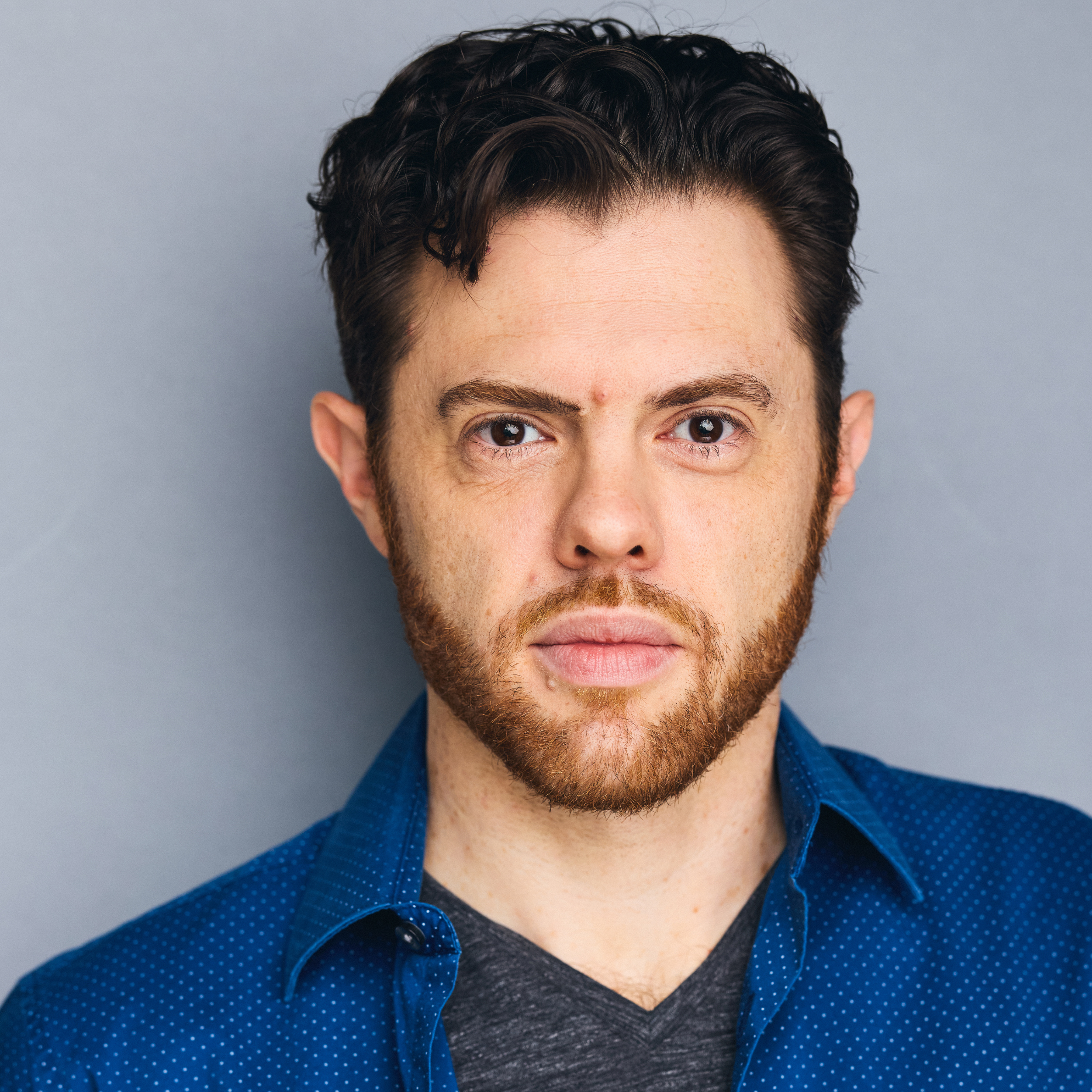 Headshot of a man with dark hair and a beard, wearing a blue button-down shirt over a gray V-neck.