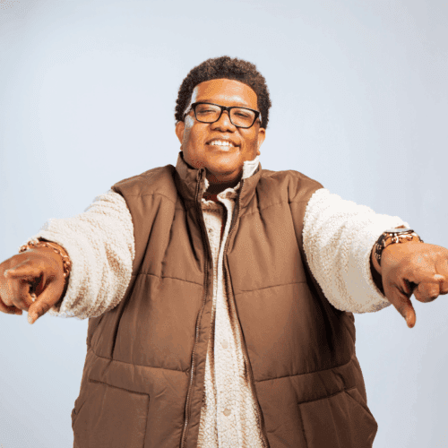 Person wearing glasses, a brown puffer vest, and a beige shirt, smiling and pointing both index fingers toward the camera against a plain light background.