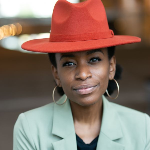 Person wearing a red wide-brim hat, gold hoop earrings, and a light green blazer, looking slightly off-camera with a neutral expression.