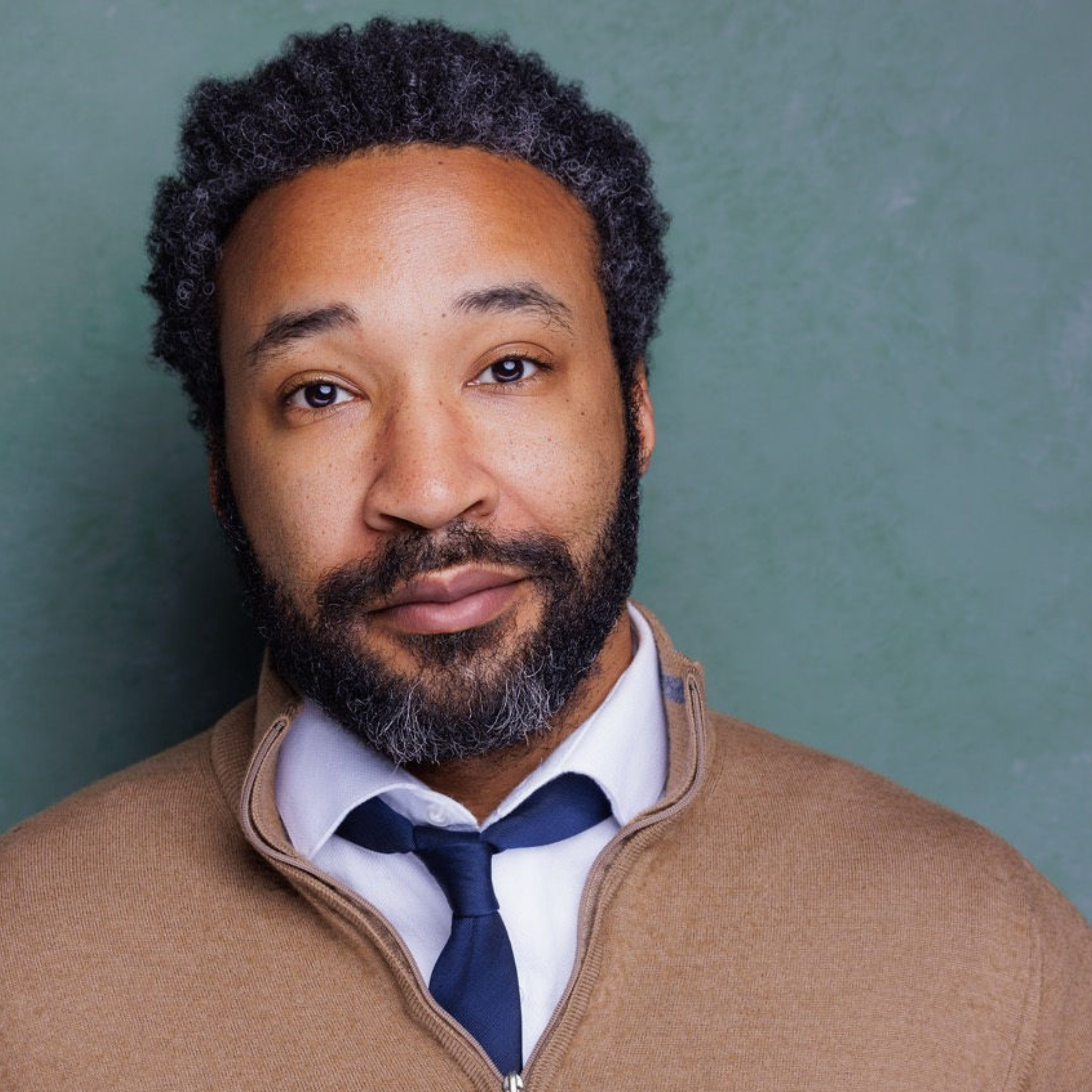 Headshot of a man with a beard, wearing a tan sweater over a white collared shirt and blue tie.