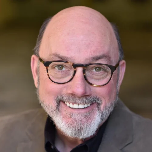 Rial Ellsworth, a middle-aged man with a bald head, gray beard, and glasses, is smiling at the camera, wearing a brown jacket and black shirt against a blurred neutral background.