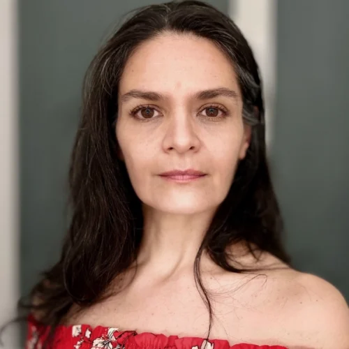 Woman with long dark hair and brown eyes wearing a red off-the-shoulder top, looking directly at the camera with a neutral expression.