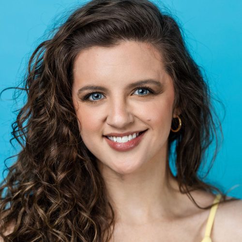 A woman with long curly brown hair and blue eyes smiles at the camera against a solid blue background. She is wearing hoop earrings and a light yellow top.