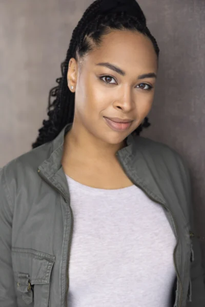 Kendra Nicole Johnson, with braided hair and a slight smile, stands against a neutral background wearing a gray jacket over a light shirt, looking confidently at the camera.