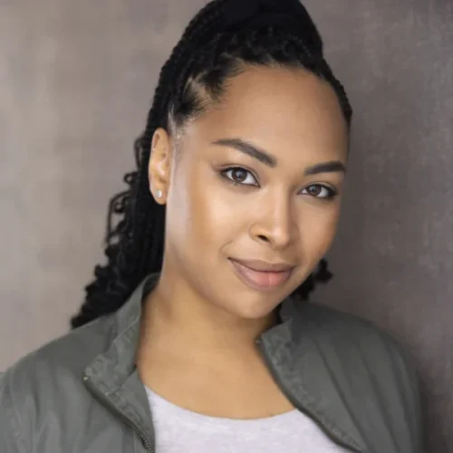Kendra Nicole Johnson, with braided hair and a slight smile, stands against a neutral background wearing a gray jacket over a light shirt, looking confidently at the camera.