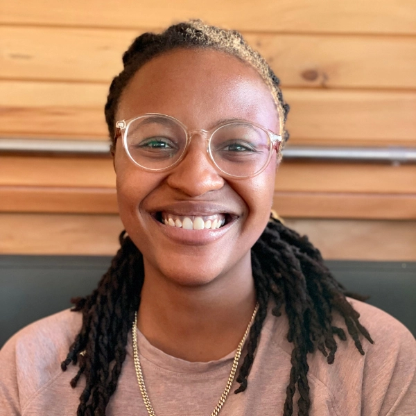 Rachael N. Blackwell, wearing glasses and a pink shirt, smiles warmly with her long dark braided hair as she sits in front of a wooden wall.