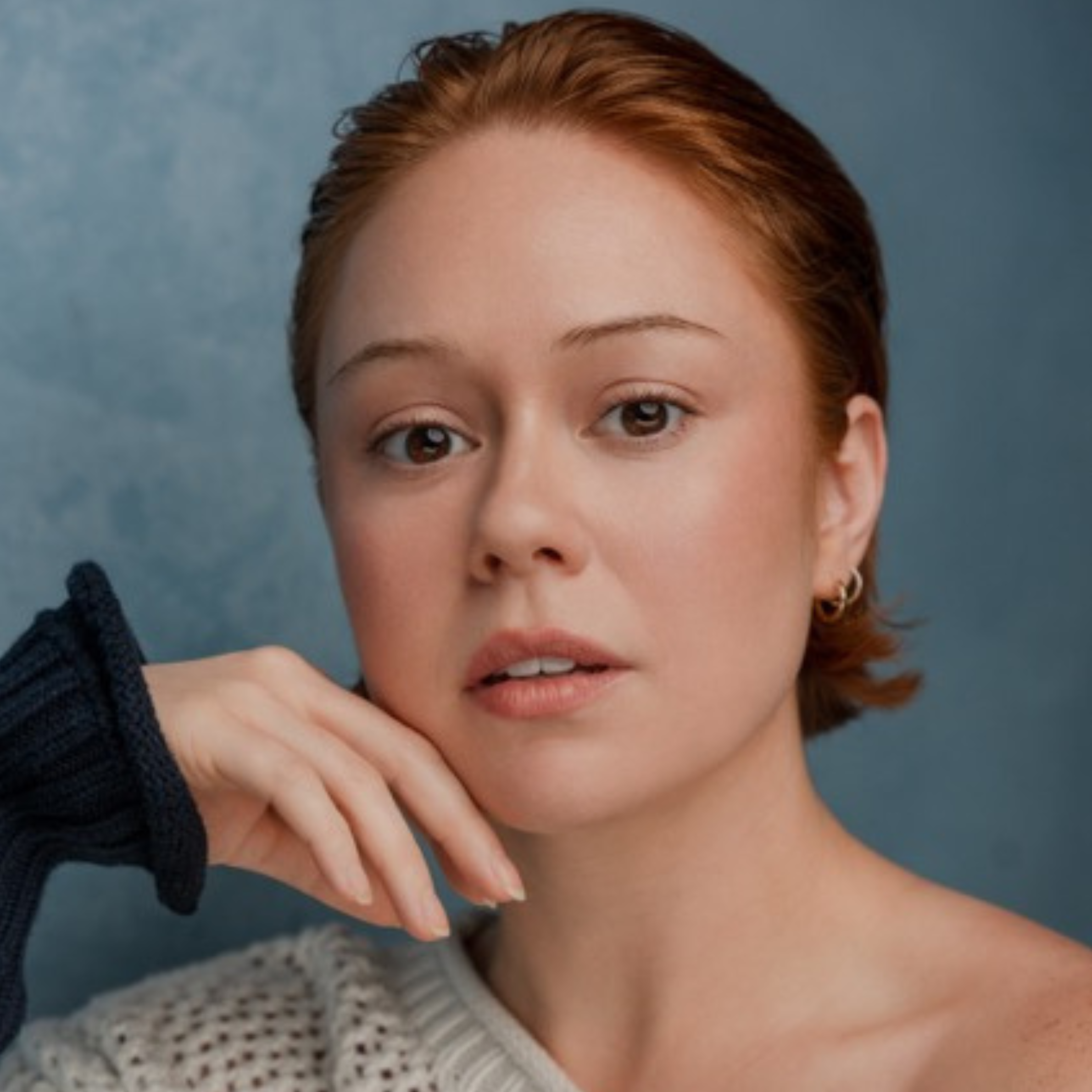 Portrait of a woman with short auburn hair resting her chin on her hand against a blue background.