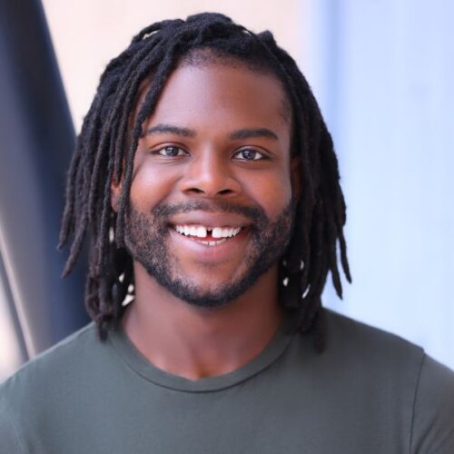 A person with medium-length dreadlocks, a short beard, and a green shirt smiles in front of a blurred blue and beige background.