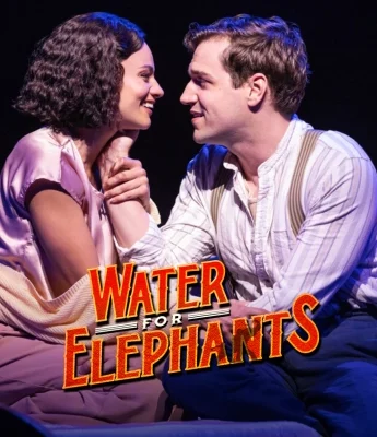 A man and woman sit close together onstage, holding hands and smiling, with the "Water for Elephants" logo in front of them.