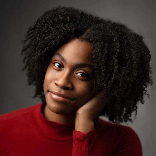 A person with curly hair wearing a red top, resting their face on their hand, looks at the camera against a dark gray background.