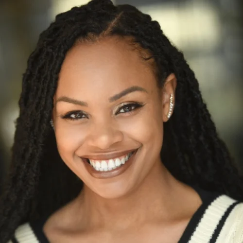 A woman with long, braided hair smiles at the camera, wearing a black and white top and multiple earrings in her left ear.