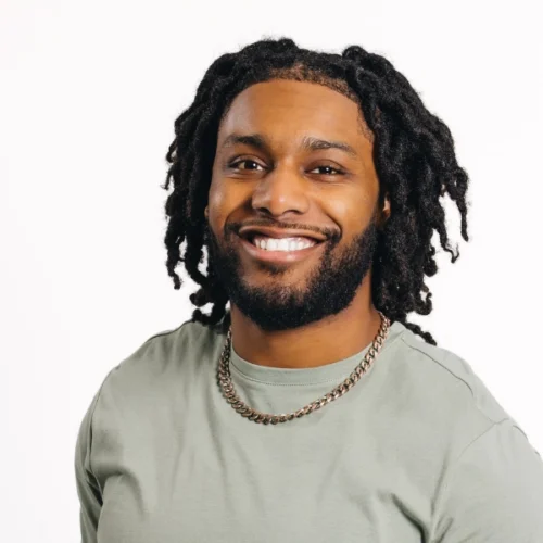A man with medium-length dreadlocks, a beard, and a light green shirt smiles at the camera against a plain white background.