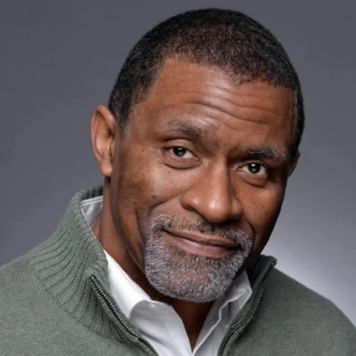 A middle-aged man with short hair and a gray beard, wearing a green zip-up sweater over a white shirt, poses against a gray background.