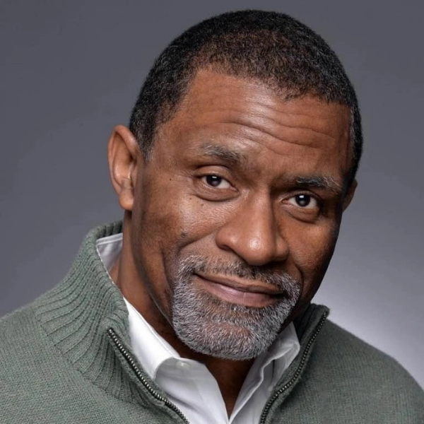 A middle-aged man with short hair and a gray beard, wearing a green zip-up sweater over a white shirt, poses against a gray background.