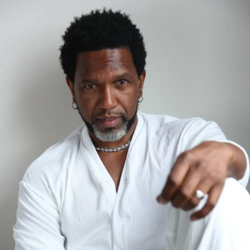 A man with short curly hair and a salt-and-pepper beard wears a white shirt and silver jewelry, sitting against a plain light background.