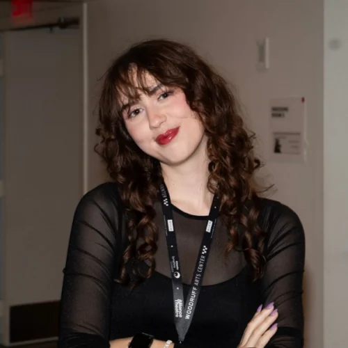 A person with long curly hair, wearing a black sheer top and a lanyard, stands indoors with arms crossed, smiling slightly at the camera.