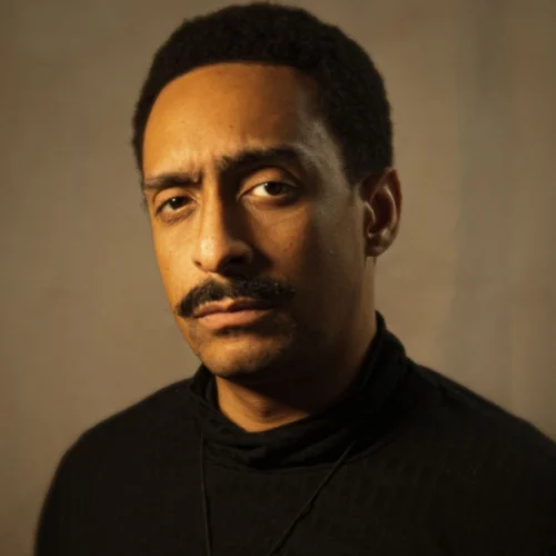 A man with short curly hair and a mustache, wearing a black turtleneck, looks toward the camera with a neutral expression against a plain background.