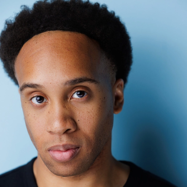 A person with short curly hair and a neutral expression stands against a light blue background, looking directly at the camera.
