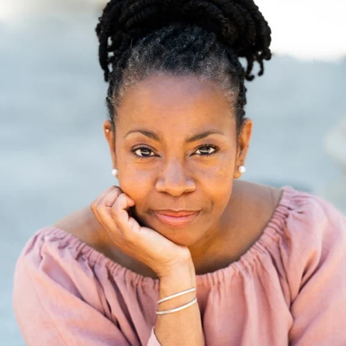 A woman with dark braided hair in a bun, wearing a pink off-the-shoulder top and pearl earrings, rests her chin on her hand and looks at the camera.