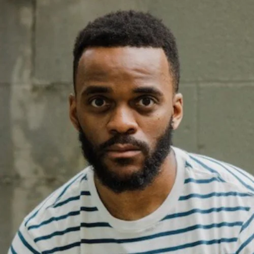 A man with short hair and a beard, wearing a white and blue striped shirt, looks directly at the camera in front of a concrete wall.