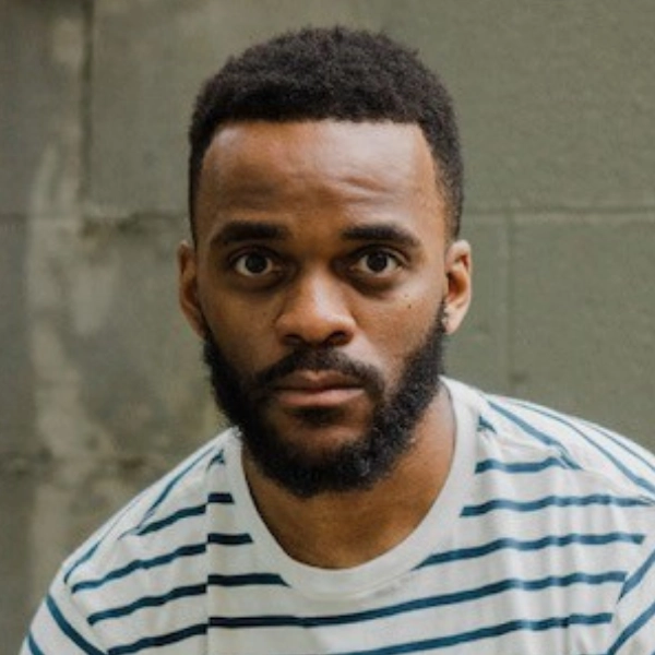 A man with short hair and a beard, wearing a white and blue striped shirt, looks directly at the camera in front of a concrete wall.