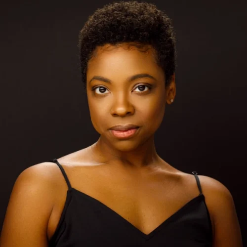 A woman with short curly hair, wearing a black spaghetti strap top, poses against a plain dark background, looking directly at the camera with a neutral expression.