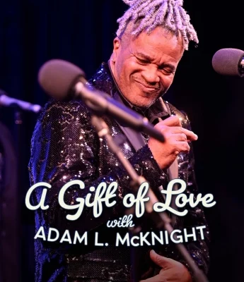 A man in a sequin jacket performs on stage, holding a microphone, with the text "A Gift of Love with Adam L. McKnight" displayed in front.