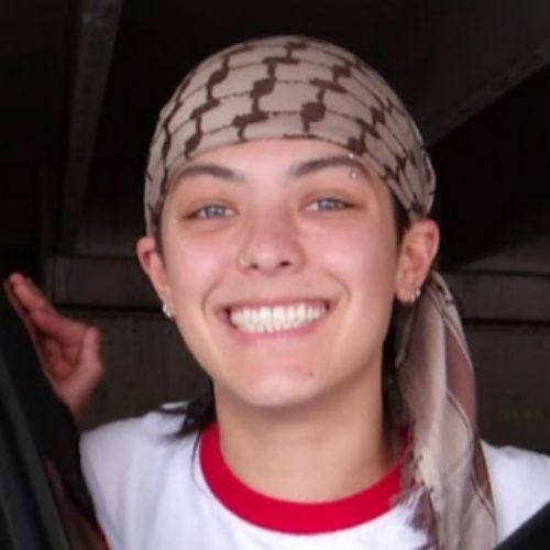 A person smiling at the camera, wearing a patterned headscarf and a white shirt with red trim, standing indoors.