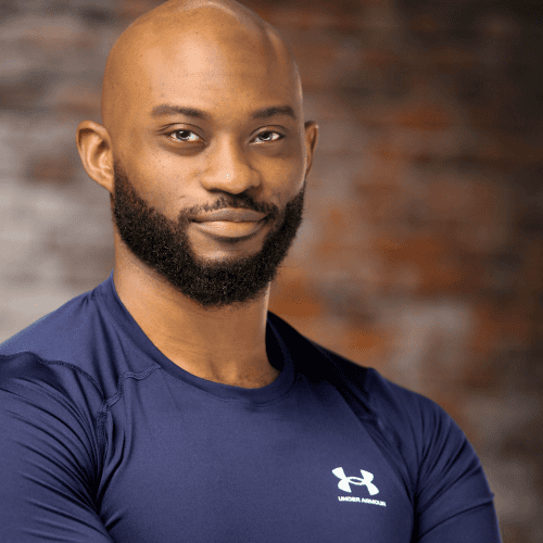 A man with a beard and shaved head wears a navy blue Under Armour shirt, standing against a blurred, brownish background, looking at the camera.
