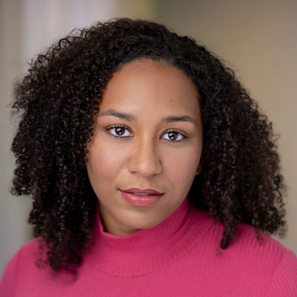 A woman with curly dark hair and brown eyes, wearing a pink turtleneck, looks directly at the camera with a neutral expression against a softly blurred background.