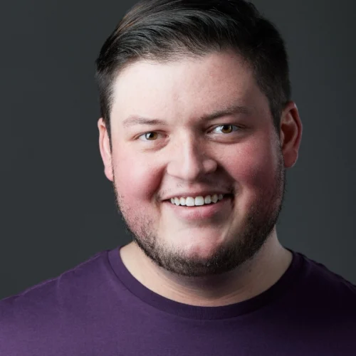 A young man with short dark hair and a trimmed beard smiles at the camera, wearing a purple shirt against a plain dark background.