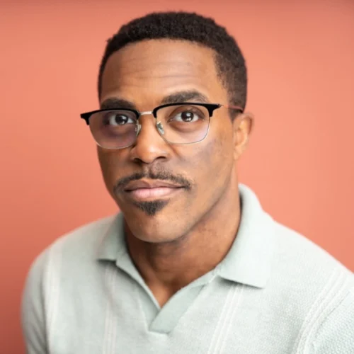 A man wearing glasses and a light green collared shirt looks at the camera, posed against a plain peach-colored background.