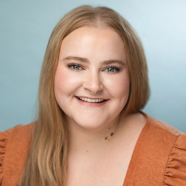 Woman with long blonde hair wearing an orange top smiles at the camera against a light blue background.