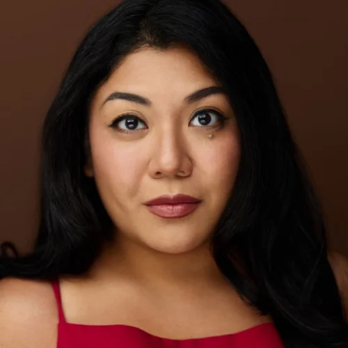 A woman with long black hair and brown eyes wears a red sleeveless top, looking directly at the camera against a brown background.