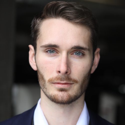 A man with short brown hair, light eyes, and a trimmed beard is wearing a dark jacket and white shirt, looking directly at the camera against a blurred background.