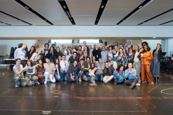 A large group of people pose together in a spacious, modern room with high ceilings and marked floors, many smiling and making playful gestures for the camera, while colorful basura bins can be seen in the background.