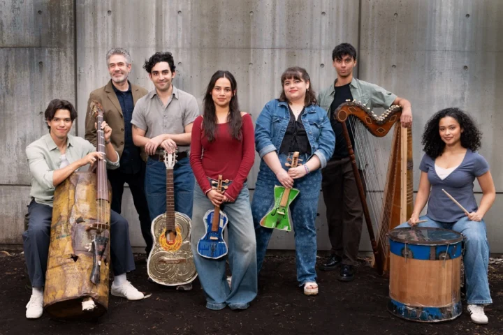 Seven people pose in front of a concrete wall, each holding a different musical instrument—strings, a harp, a drum, and even a large wooden Basura instrument add unique flair to the group.