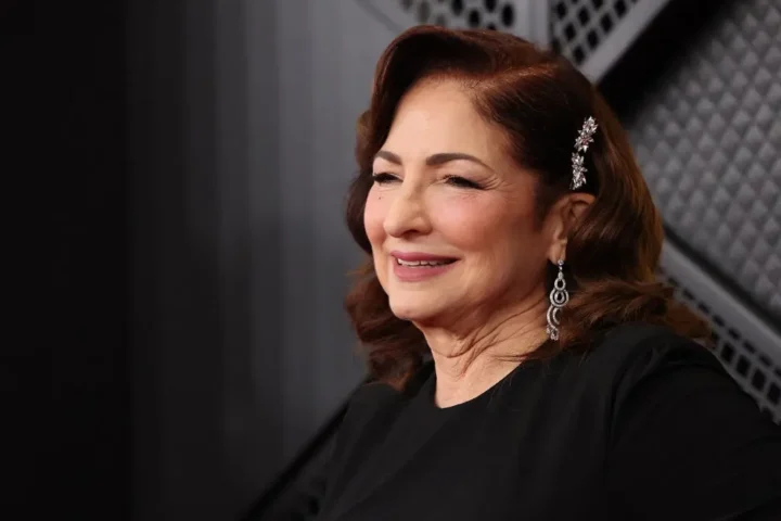 A woman with wavy brown hair, wearing a black top, jeweled hairpin, and dangling earrings, smiles while posing in front of a patterned Basura-themed background.